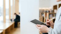 Businessman in light gray shirt holding tablet device in modern office environment with blurred colleagues and wooden interior details in background.
