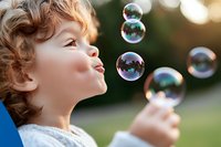 Joyful Caucasian child with curly hair blowing soap bubbles outdoors in summer garden. Natural bokeh background creates magical atmosphere for childhood memories.