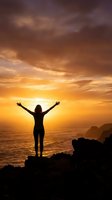 Woman silhouette with raised arms celebrating freedom at golden sunset over ocean waves from rocky cliff for wellness and motivation concepts.