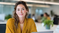 Young professional woman in mustard yellow blouse smiling confidently in modern office environment with blurred colleagues in background.