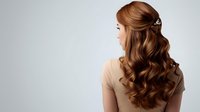 Young woman with long wavy brown hair styled in elegant curls wearing beige top against light background, view from behind, showcasing professional hairstyling.