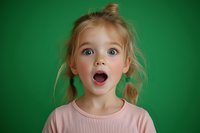 Young girl with blonde pigtails expressing surprise and wonder, mouth open wide with amazement, wearing pink shirt against green background, capturing pure childhood emotion.