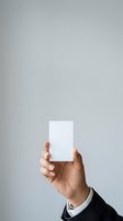 Cropped view of male hand with medium skin tone holding blank white card against neutral gray background, wearing dark sleeve with white cuff for business, marketing, or branding mockup.