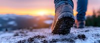Hiking boot step in snow at sunset panorama, close-up view of winter trekking footwear against mountain landscape and orange sky background.