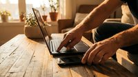 Person working on laptop computer at wooden desk in warm sunlight creating cozy home office atmosphere for remote work and productivity.