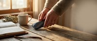 Male hands using smartphone at wooden desk with coffee cup and newspaper in warm morning sunlight for communication and daily routine.