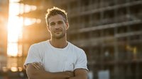 Confident young Caucasian man in white t-shirt with crossed arms smiling at camera during golden hour sunset with urban building background for lifestyle content.