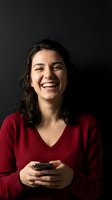 Happy young Hispanic woman laughing while using smartphone wearing red sweater against dark background for lifestyle and technology concepts.