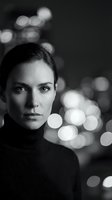 Young Caucasian woman in black turtleneck sweater with intense gaze against blurred city lights bokeh background in monochrome portrait photography.