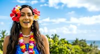 Young woman with long dark hair and warm skin tone wearing vibrant tropical flower lei and colorful floral garland, smiling outdoors with ocean and palm trees in background.
