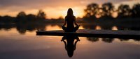 Woman meditating in lotus pose on wooden dock at sunset with calm lake reflection creating peaceful zen atmosphere for wellness and mindfulness practice.