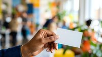 Close-up of hand holding blank white business card with blurred office environment and colleagues in background, suitable for corporate branding and professional networking.