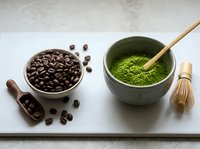 Coffee beans in wooden bowl next to matcha green tea powder with bamboo whisk on marble surface for beverage preparation and culinary arts.