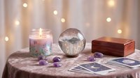Mystical fortune telling setup with crystal ball, tarot cards, candle and amethyst stones on velvet cloth with warm bokeh lights creating magical atmosphere.