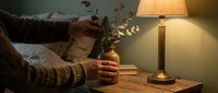 Cozy evening scene with person in knit sweater holding small vase with eucalyptus branches beside warm table lamp on wooden surface creating hygge atmosphere.