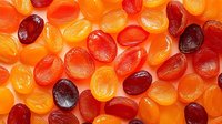 Colorful assorted fruit gummy candies in red, orange and yellow hues scattered on bright orange background for confectionery and sweet treat designs.
