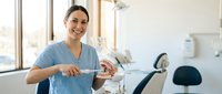 Young Hispanic female dentist in blue scrubs smiling while holding dental tools in modern clinic office with equipment and natural lighting for healthcare marketing.