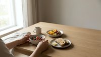 Hands reaching for healthy breakfast bowl on wooden table with coffee, fruit and pastries in bright morning light streaming through window.