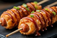 Crispy corn dogs on wooden skewers drizzled with ketchup and mustard sauces, garnished with fresh chopped green onions, served on black slate surface.