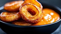 Golden crispy onion rings with caramelized coating served alongside tangy orange dipping sauce in dark bowl for restaurant menu and comfort food concepts.