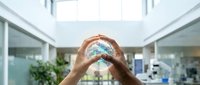 Young adult with medium skin tone forming heart shape with hands around globe, symbolizing environmental care and sustainability in modern office setting.