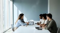 Professional businesswoman presenting financial data to diverse team in modern conference room with natural lighting for corporate meetings.