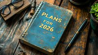 Blue vintage notebook with Plans for 2026 text on wooden desk surrounded by glasses, pen and office supplies for goal setting and planning concepts.