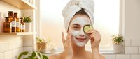 Young woman applying cucumber slice facial mask in modern bathroom with towel on head for natural skincare routine and spa treatment at home.