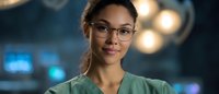 Young African American female medical professional wearing glasses and scrubs smiles confidently in hospital setting with bokeh lights in background.