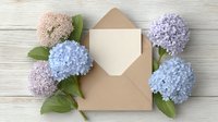 Pastel hydrangea flowers frame open kraft paper envelope on white wooden background. Fresh spring floral composition for wedding invitation or greeting card design.