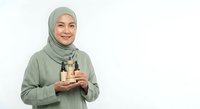 Young Muslim woman in green hijab holding skincare bottles on white background for beauty and cosmetics marketing campaigns.