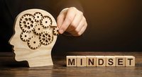 Business professional adjusting wooden gears in human head silhouette representing mindset development and mental growth with letter blocks on desk.