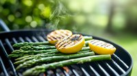 Grilled lemon halves and fresh asparagus spears on barbecue grate with rising smoke, creating summer outdoor cooking atmosphere against blurred green background.