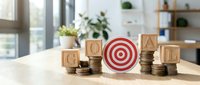 Wooden blocks spelling GOAL with target symbol and coin stacks on desk representing financial planning and achievement strategy for business success.