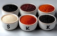 Colorful assortment of fresh caviar in white ceramic bowls with black lettering, including red, black and golden varieties arranged in two rows on gray surface.