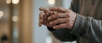 Senior man's weathered hands clasped together in prayer or contemplation, showing age and wisdom through gentle gesture in warm indoor setting.