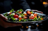 Fresh mixed greens salad with cherry tomatoes and cheese served on dark plate by hands in restaurant setting with warm bokeh lighting background.