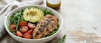 Grilled chicken breast sliced with couscous, fresh tomatoes, arugula and avocado in white bowl on wooden surface for healthy lunch or dinner meal preparation.