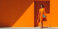 Stylish woman in vibrant orange outfit walking confidently against matching orange wall with dramatic shadow, carrying shopping bags and blue clutch purse.