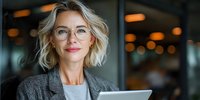 Confident middle-aged businesswoman with glasses and blonde wavy hair holding digital tablet in modern office environment.