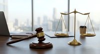 Legal office desk with wooden gavel and golden scales of justice near laptop computer in modern law firm with city skyline background for attorney services.