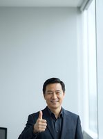 Middle-aged Asian male professional wearing dark blazer and navy shirt giving thumbs up gesture in bright modern office with white walls and natural window light, expressing approval and success.