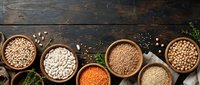 Assorted legumes and grains in wooden bowls on dark rustic surface, overhead view for food styling, cooking, nutrition, and recipe content.