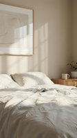 Serene bedroom with unmade bed, white linens, natural sunlight streaming through window, minimalist decor, wooden nightstand with mug and plant, framed artwork on wall.