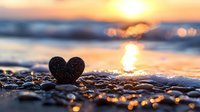 Heart-shaped stone silhouette on beach pebbles at golden sunset with ocean waves and sun reflection creating romantic bokeh background for Valentine's Day cards.