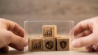 Business hands protecting wooden blocks with growth chart, pie chart and shield icons representing financial security and data protection strategies.