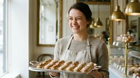 Smiling female baker with dark hair and South Asian ethnicity wearing beige apron holds fresh baked croissants in bakery shop with golden lighting and display cases.