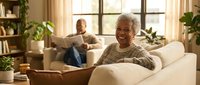 Happy senior African American woman relaxing on sofa at home with husband reading in background, cozy living room interior with plants and natural light.