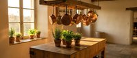 Rustic kitchen interior with copper pots hanging from wooden rack above butcher block island. Herb plants on windowsill create cozy farmhouse cooking space.