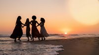 Four women holding hands dancing in circle silhouette on beach at sunset with golden sky and ocean waves creating joyful friendship moment during summer vacation.
